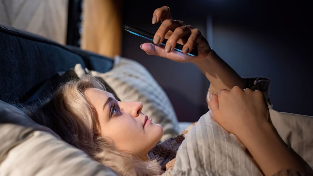 Jovem acordada na cama utilizando o celular próximo ao rosto